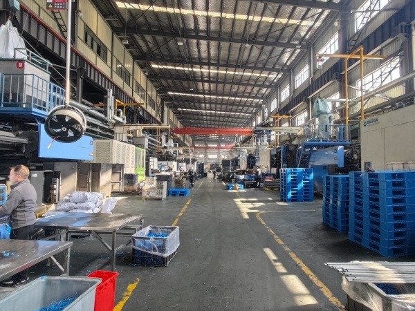An expansive factory interior with large machinery, blue plastic pallets neatly stacked, and workers at stations, indicating an active plastic pallet production environment.