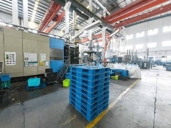 An industrial factory interior with large machinery, red overhead cranes, and stacks of blue plastic pallets. It shows an active production environment for plastic pallets.