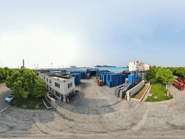 An aerial view of an industrial site with blue plastic pallets stacked outside buildings. Trucks and greenery are visible, indicating a logistics and production area for plastic pallets.