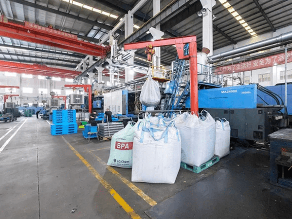 An industrial factory scene with large machinery, blue plastic pallets, and bulk bags labeled "BPA". It reflects the production environment of plastic pallet manufacturing.