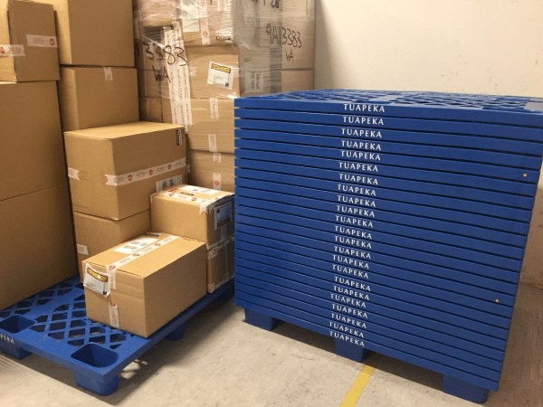 In a storage area, blue plastic pallets branded "TUAPEKA" are used to stack and organize cardboard boxes, demonstrating their practical application in warehousing.