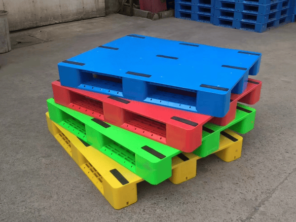 A stack of colorful plastic pallets in blue, red, green, and yellow, neatly arranged on a concrete floor, showcasing their vibrant hues and robust design for industrial use.