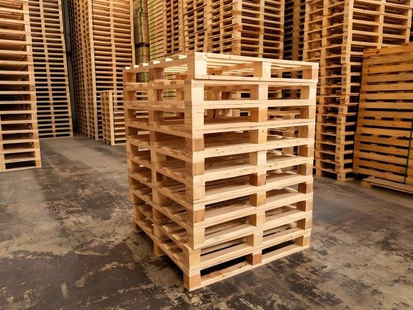 A stack of neatly arranged light - brown wooden pallets in the center, with more in the background in a warehouse. The gray floor shows signs of wear, presenting an industrial storage scene. 