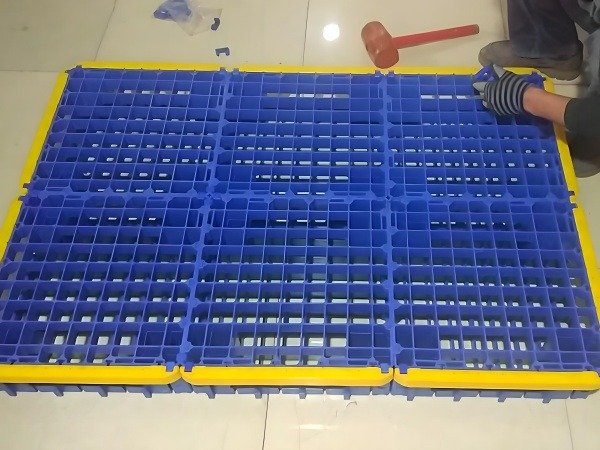 A person wearing dark clothes and gloves is assembling or repairing a blue plastic splice pallet with yellow edges using red tools on a light colored floor scattered with small items.