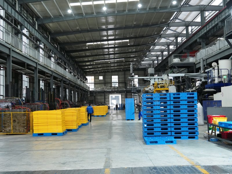 The interior of a spacious factory with high ceilings, where yellow and blue plastic pallets are stacked. A worker is visible in the distance, indicating ongoing industrial activity.