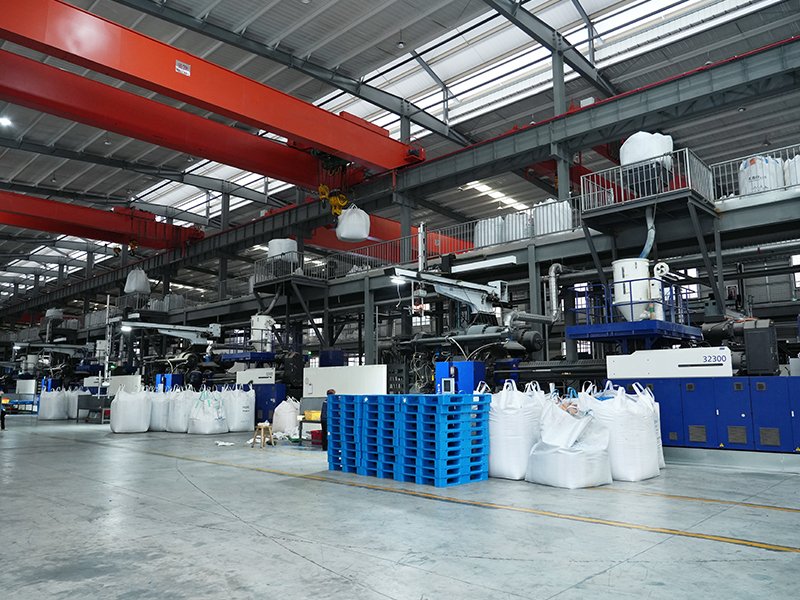 An industrial factory interior with large red overhead cranes, various machinery, and numerous white bulk bags and blue pallets on the floor.
