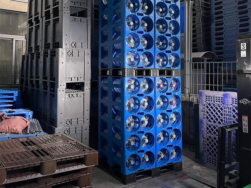 A stack of blue plastic water racks filled with large water bottles, situated in an industrial or storage area with other pallets and equipment around.
