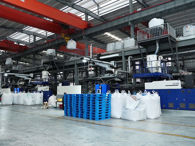 An industrial factory interior with large machinery and red overhead cranes. Blue plastic pallets are placed alongside large white bulk bags, indicating a manufacturing or production environment for plastic pallets.