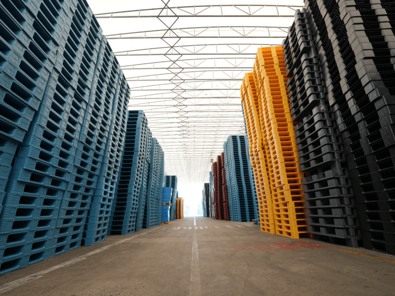 Double sided pallets in yellow, green, black, blue, and red, as well as nine legged pallets, are stacked high and placed in the factory.