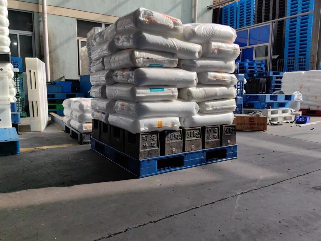 In an industrial area, a blue 3 - runners pallet bears stacks of white packaged goods, potentially relevant to 3 runners pallet load testing, showing its load - bearing capacity.