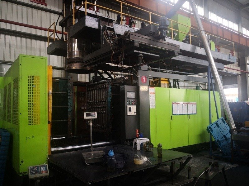 A large - scale injection molding machine in a factory, used for manufacturing plastic pallets. It features a control panel, metal components, and is situated in a spacious industrial setting.