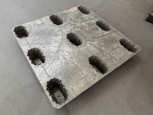 A gray plastic pallet with a textured surface and multiple hollow sections is placed on a gray floor, highlighting its design for industrial applications.
