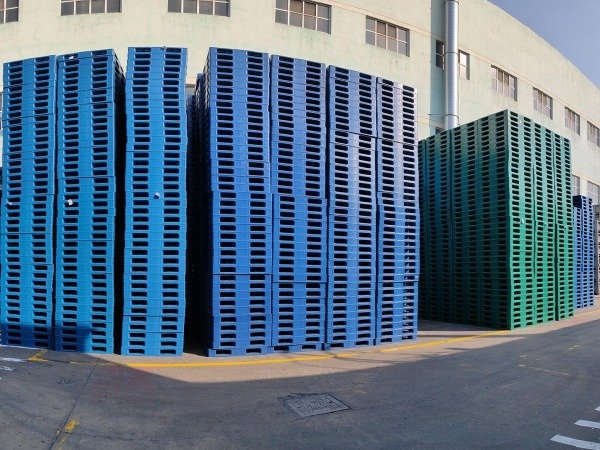 Stacks of blue and green plastic pallets outside a factory. In Singapore, standard pallet sizes include 1100mm×1100mm, 1200mm×1000mm etc., used in logistics and transport.