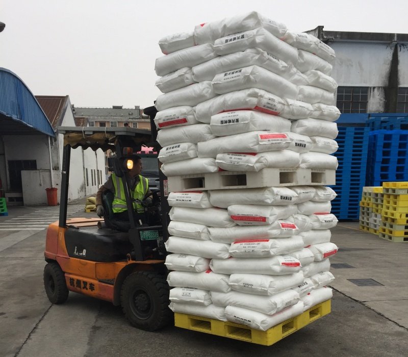 Forklift carrying white sacks stacked on a yellow pallet in an industrial area.
