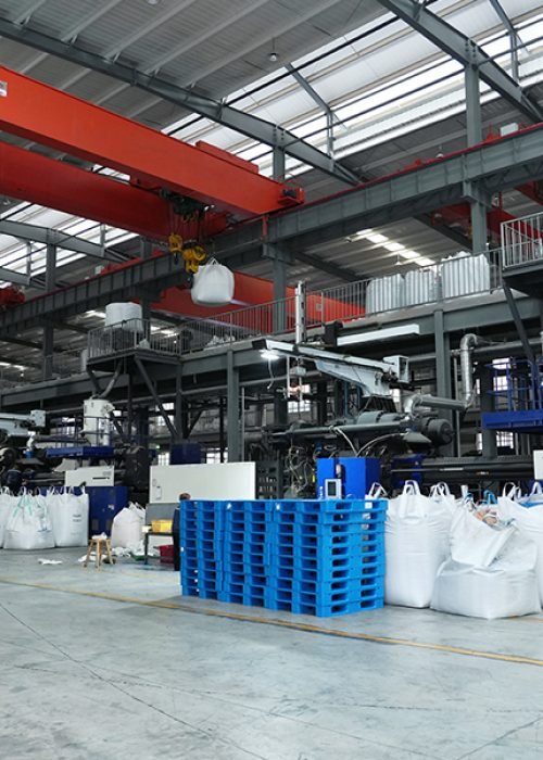 An industrial factory interior with large red overhead cranes, various machinery, and numerous white bulk bags and blue pallets on the floor.