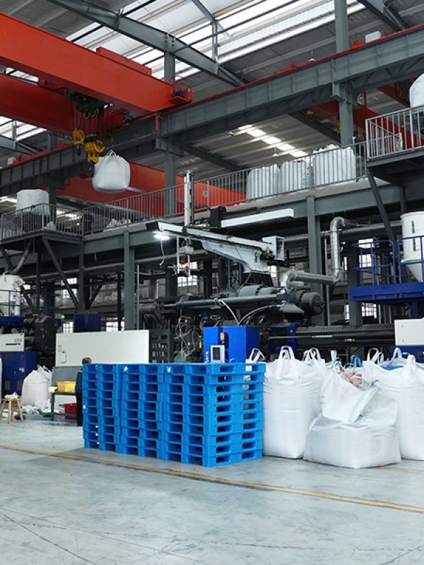 An industrial factory interior with large machinery and red overhead cranes. Blue plastic pallets are placed alongside large white bulk bags, indicating a manufacturing or production environment for plastic pallets.